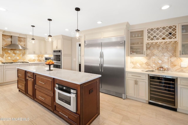 kitchen with stainless steel appliances, decorative light fixtures, cream cabinetry, wine cooler, and wall chimney range hood