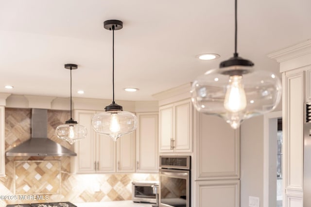 kitchen featuring cream cabinets, oven, wall chimney range hood, decorative backsplash, and pendant lighting