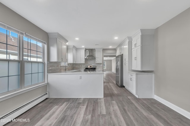 kitchen with stainless steel refrigerator, white cabinetry, baseboard heating, kitchen peninsula, and wall chimney exhaust hood