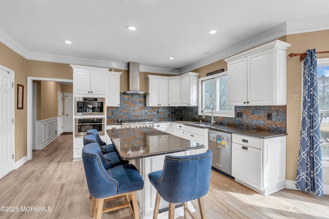 kitchen with stainless steel appliances, a sink, a kitchen island, white cabinets, and wall chimney range hood