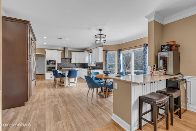 kitchen featuring a breakfast bar area, a peninsula, white cabinetry, hanging light fixtures, and wall chimney range hood