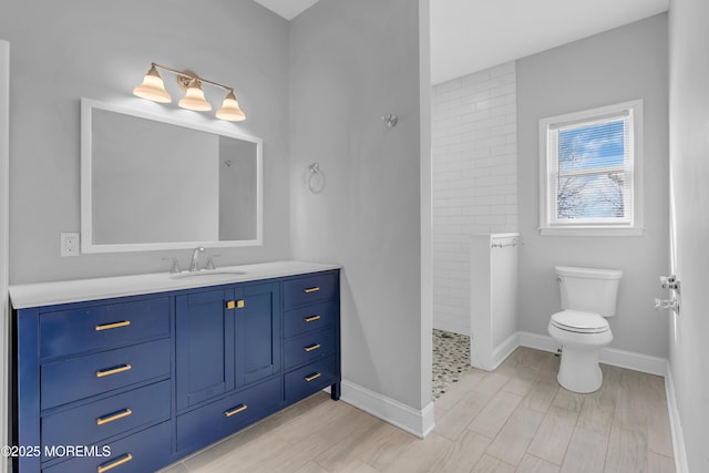 bathroom with toilet, wood finished floors, vanity, baseboards, and a tile shower