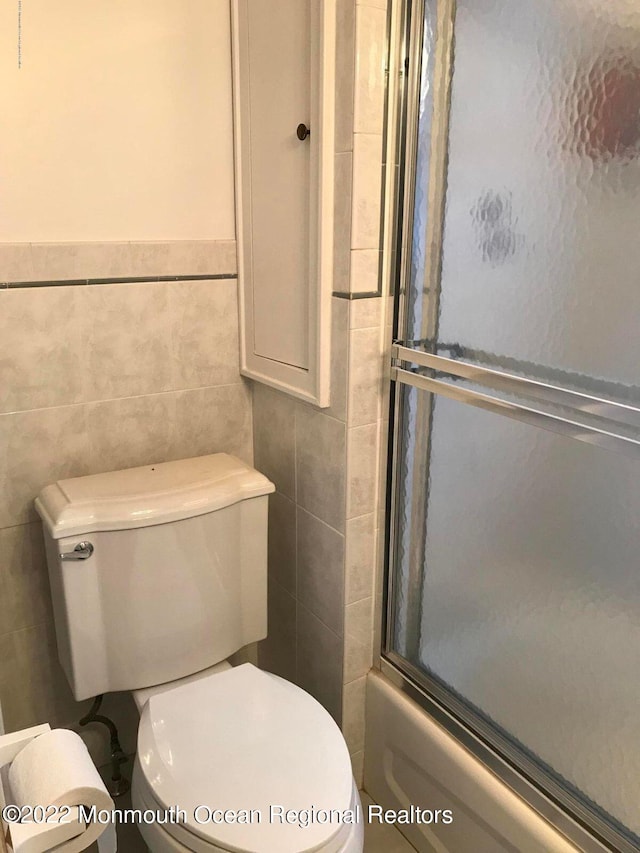 bathroom featuring tile walls, shower / bath combination with glass door, and toilet