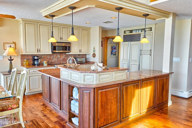 kitchen with a baseboard radiator, a kitchen island with sink, stainless steel microwave, and cream cabinetry