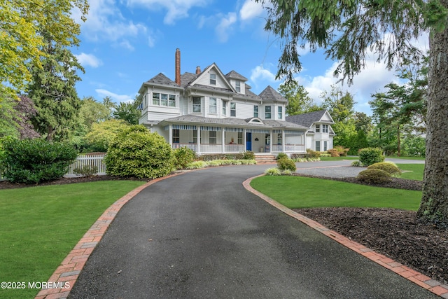 view of front of house with aphalt driveway, a chimney, a porch, fence, and a front lawn