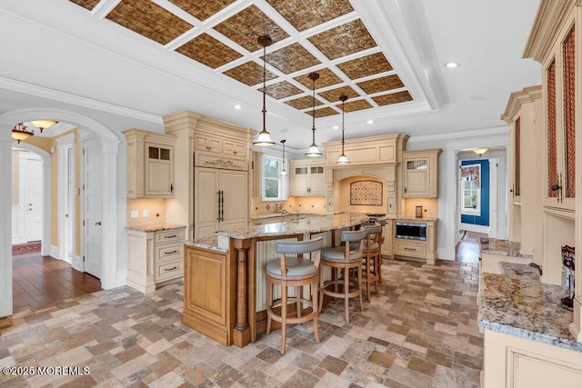 kitchen with arched walkways, a breakfast bar area, glass insert cabinets, coffered ceiling, and a large island with sink