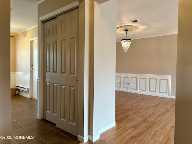 corridor with a notable chandelier, a baseboard heating unit, a wainscoted wall, visible vents, and crown molding