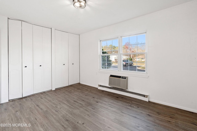 unfurnished bedroom with a baseboard radiator, hardwood / wood-style flooring, a wall mounted AC, and two closets