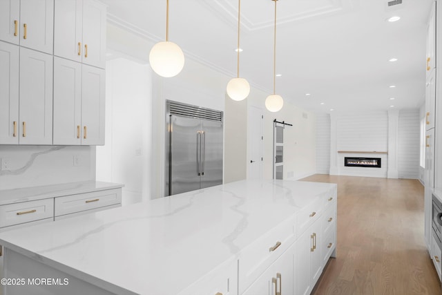 kitchen with light stone counters, built in refrigerator, white cabinets, a barn door, and hanging light fixtures