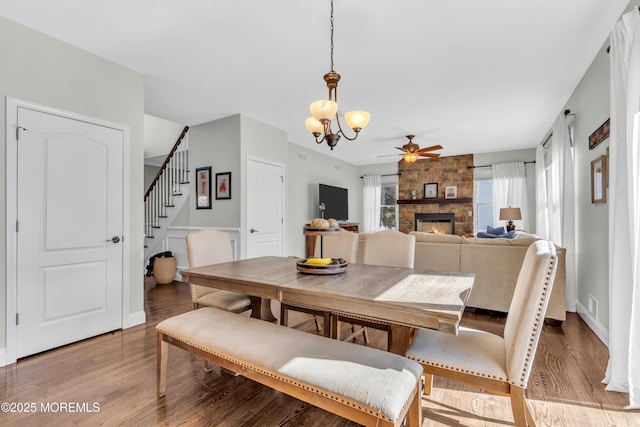 dining area with stairs, a stone fireplace, ceiling fan with notable chandelier, and light wood-style flooring