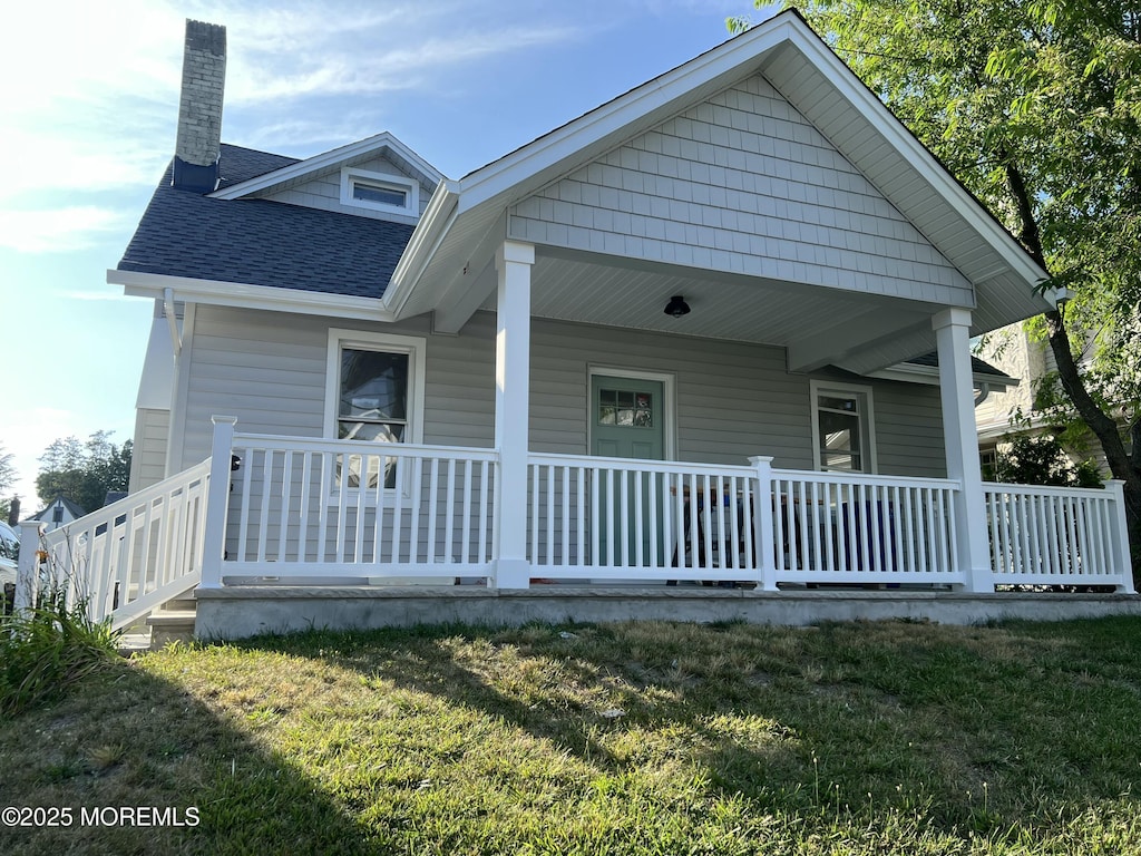 rear view of property with covered porch, roof with shingles, a yard, and a chimney