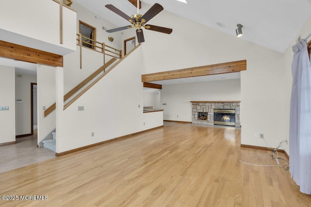 unfurnished living room with stairway, a ceiling fan, a stone fireplace, light wood-type flooring, and baseboards