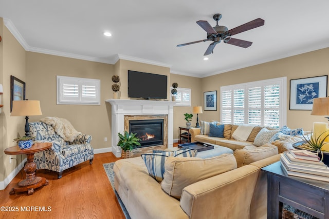 living room featuring crown molding, light wood-style flooring, a ceiling fan, a glass covered fireplace, and baseboards