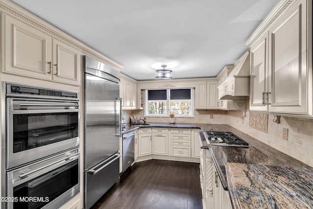kitchen featuring dark stone counters, appliances with stainless steel finishes, wall chimney range hood, and cream cabinetry