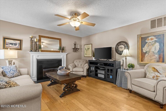 living room with light wood finished floors, visible vents, a glass covered fireplace, ceiling fan, and a textured ceiling