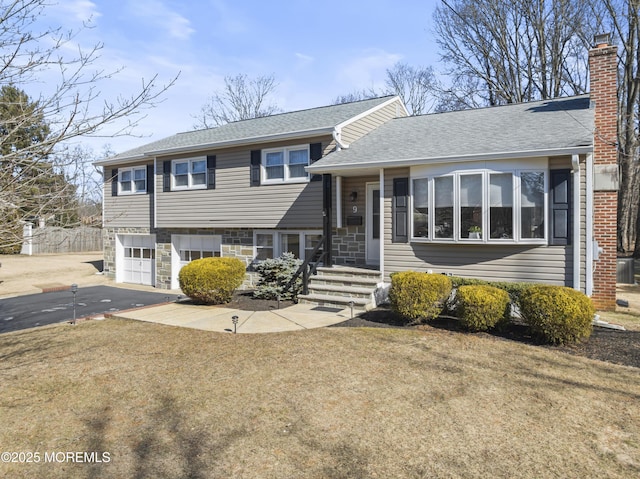 split level home with driveway, a garage, a shingled roof, stone siding, and a chimney