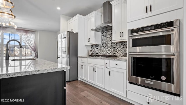 kitchen with light stone counters, stainless steel appliances, a sink, white cabinets, and wall chimney range hood