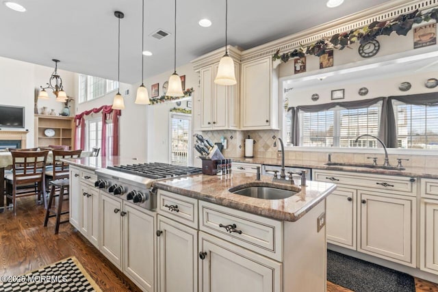 kitchen featuring decorative light fixtures, stainless steel gas cooktop, a center island with sink, a sink, and light stone countertops