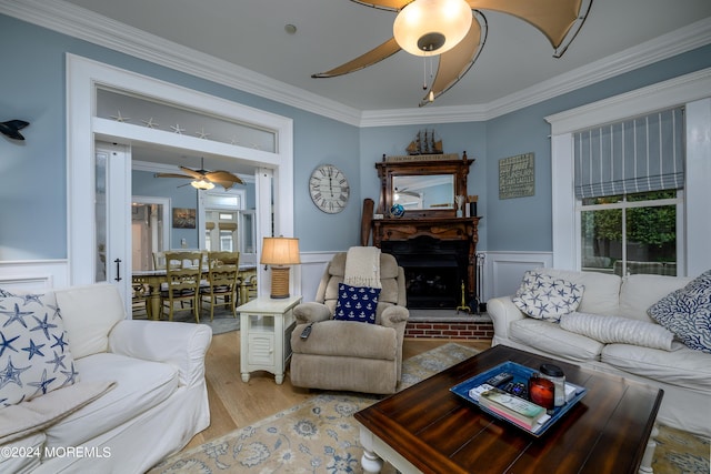 living room featuring a wainscoted wall, crown molding, a fireplace, ceiling fan, and light wood-type flooring