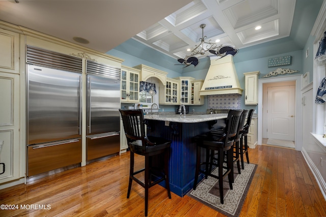 kitchen with cream cabinets, glass insert cabinets, decorative light fixtures, stainless steel built in refrigerator, and a center island with sink