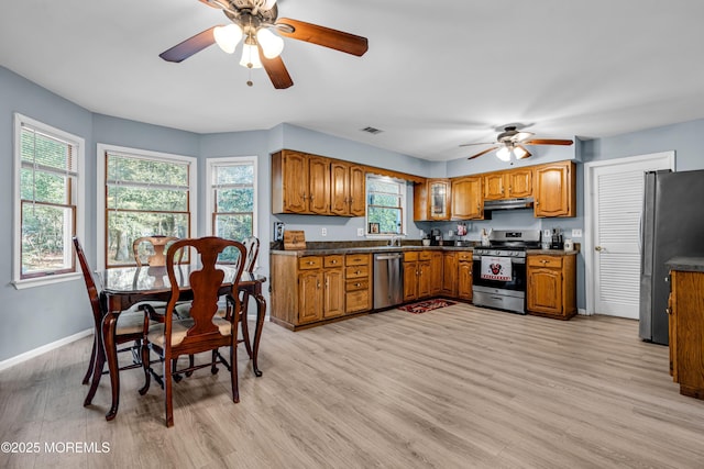 kitchen with brown cabinets, stainless steel appliances, dark countertops, light wood-style flooring, and under cabinet range hood