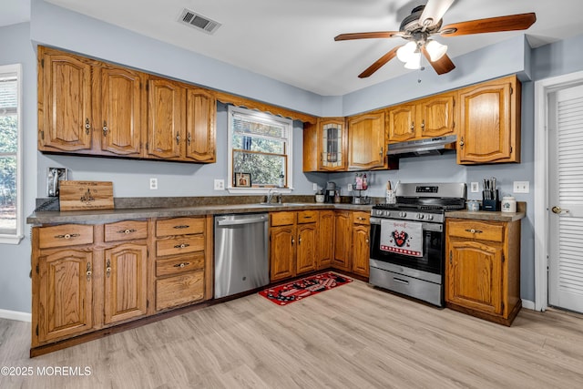 kitchen with brown cabinetry, dark countertops, light wood-style flooring, appliances with stainless steel finishes, and under cabinet range hood