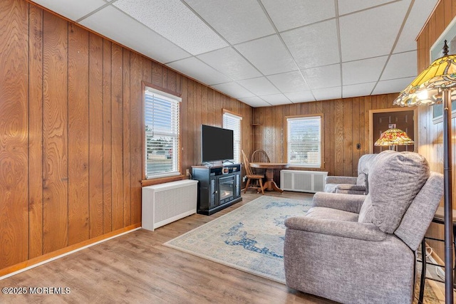 living room featuring a drop ceiling, wood walls, radiator heating unit, wood finished floors, and a glass covered fireplace