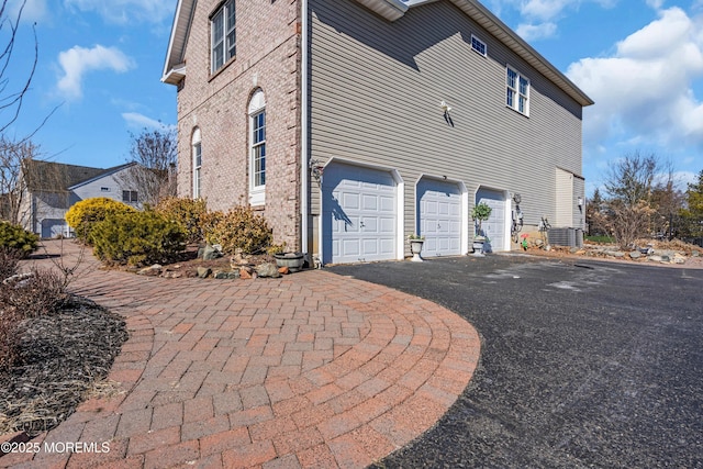 view of property exterior featuring driveway, an attached garage, central AC, and brick siding