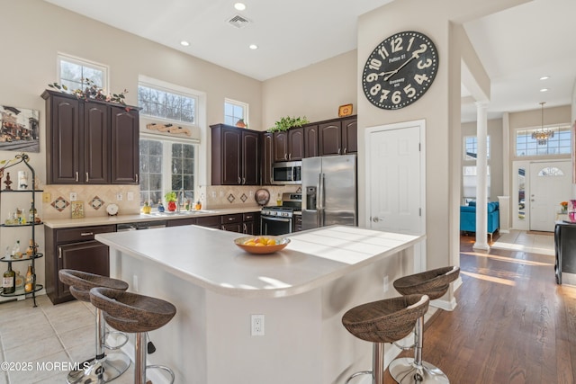 kitchen featuring dark brown cabinetry, appliances with stainless steel finishes, a center island, ornate columns, and a kitchen bar