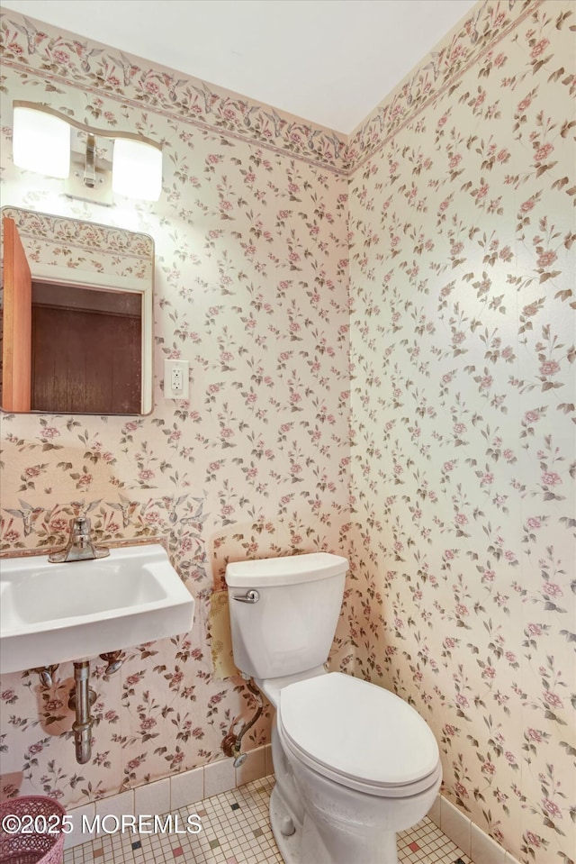 bathroom featuring baseboards, wallpapered walls, a sink, tile patterned flooring, and toilet