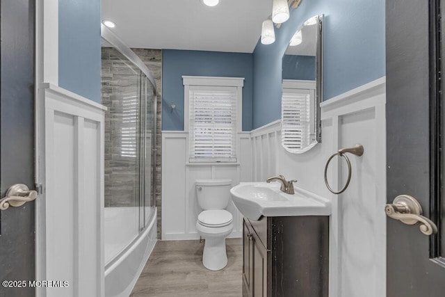 bathroom with toilet, wainscoting, combined bath / shower with glass door, vanity, and wood finished floors