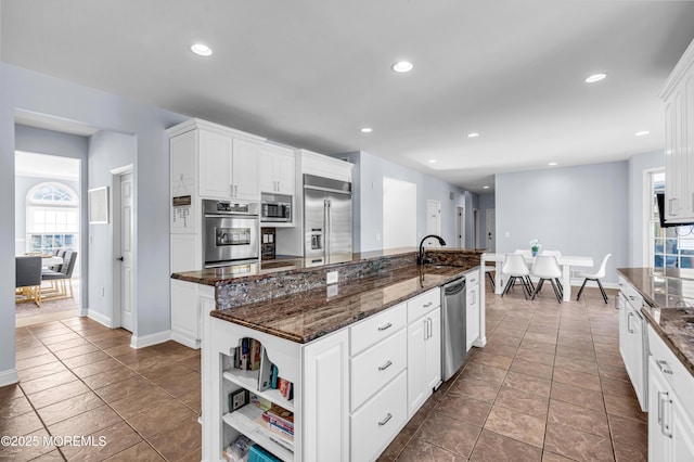 kitchen featuring dark stone counters, built in appliances, a kitchen island with sink, white cabinetry, and recessed lighting