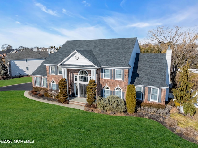 neoclassical home featuring brick siding, driveway, roof with shingles, a chimney, and a front yard