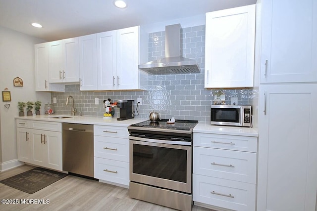 kitchen with stainless steel appliances, light countertops, wall chimney range hood, white cabinetry, and a sink