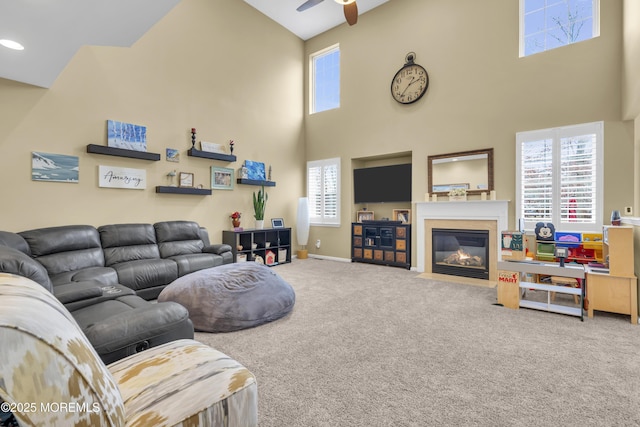 living area featuring baseboards, a towering ceiling, ceiling fan, a fireplace with flush hearth, and carpet