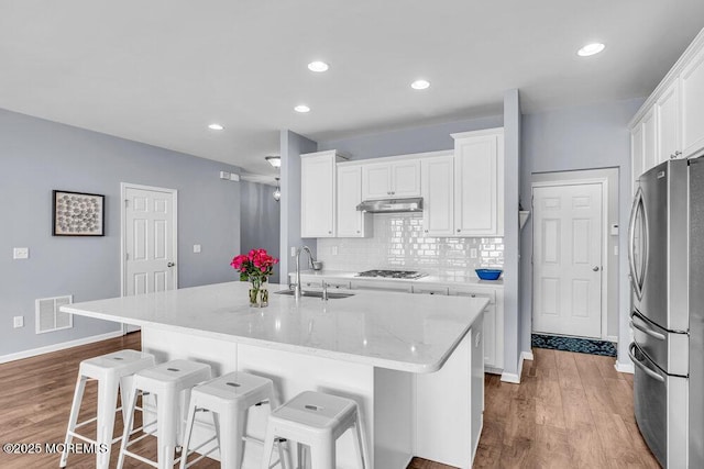 kitchen with under cabinet range hood, a sink, white cabinets, backsplash, and freestanding refrigerator