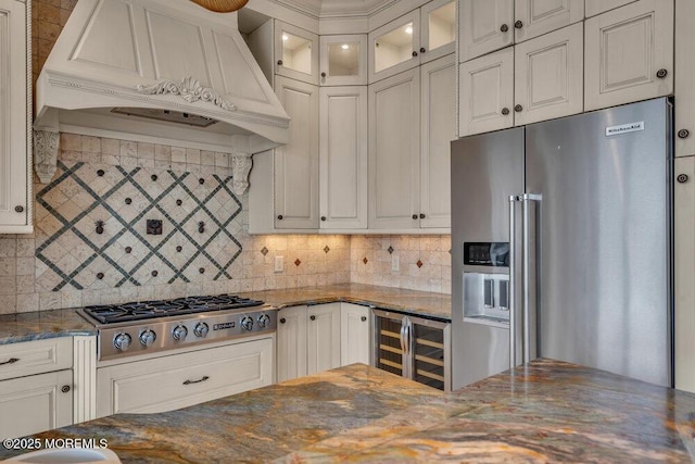 kitchen with wine cooler, custom exhaust hood, stainless steel appliances, glass insert cabinets, and dark stone counters