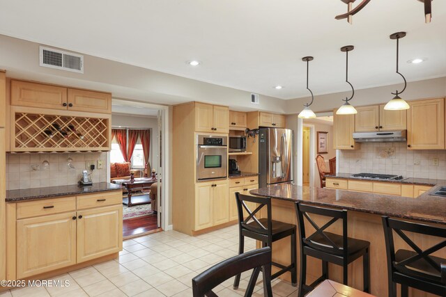 kitchen featuring light brown cabinets, under cabinet range hood, visible vents, a kitchen breakfast bar, and appliances with stainless steel finishes