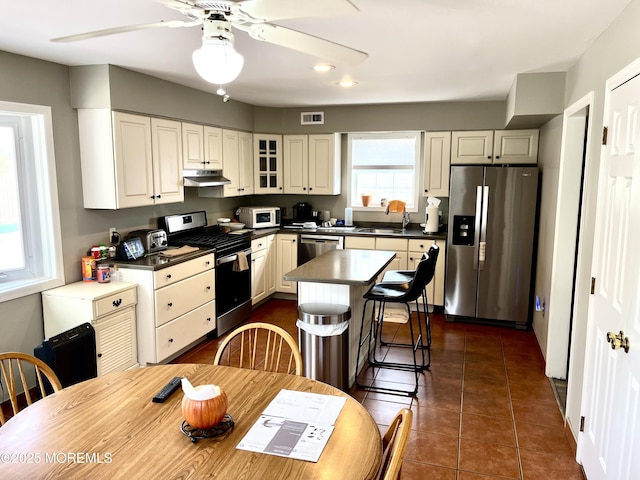 kitchen with under cabinet range hood, visible vents, appliances with stainless steel finishes, dark countertops, and glass insert cabinets