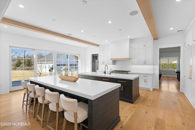 kitchen featuring light wood finished floors, premium range hood, a kitchen island with sink, and beam ceiling