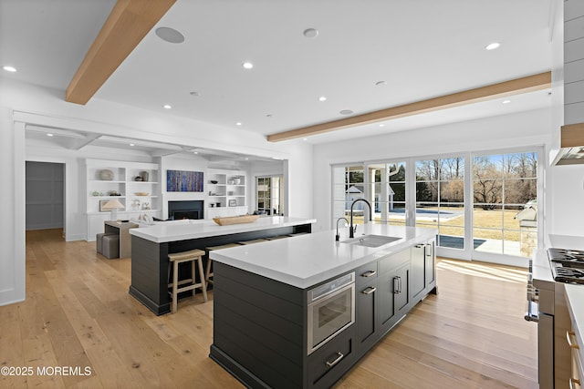 kitchen featuring light wood-type flooring, stainless steel microwave, beamed ceiling, and an island with sink