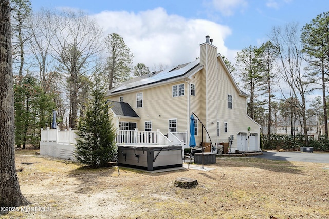 rear view of property featuring a garage, a wooden deck, solar panels, a chimney, and a hot tub