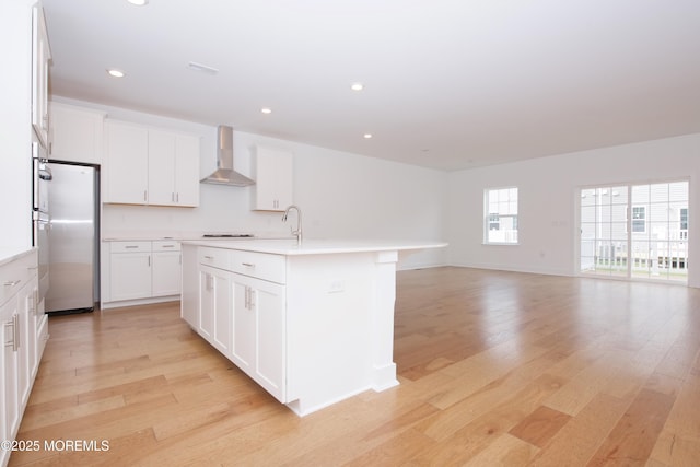 kitchen featuring wall chimney range hood, a kitchen island with sink, light wood finished floors, and freestanding refrigerator