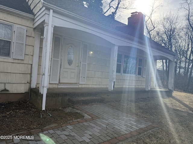 entrance to property with covered porch, a chimney, and roof with shingles