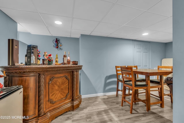 bar with a bar, light wood-type flooring, a drop ceiling, and baseboards