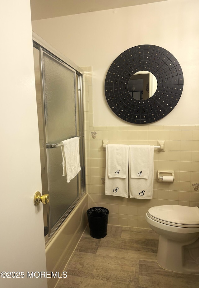 bathroom featuring wainscoting, toilet, wood finished floors, combined bath / shower with glass door, and tile walls