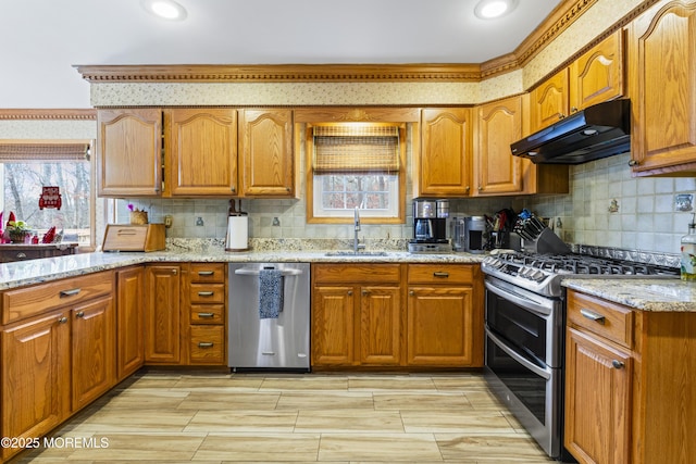 kitchen featuring brown cabinets, appliances with stainless steel finishes, a sink, light stone countertops, and under cabinet range hood