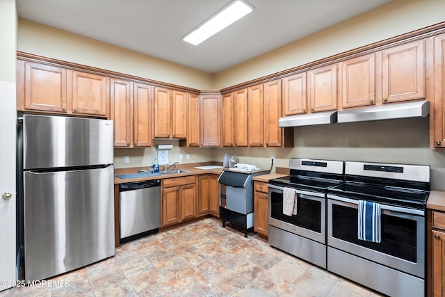 kitchen with light countertops, appliances with stainless steel finishes, stone finish flooring, a sink, and under cabinet range hood