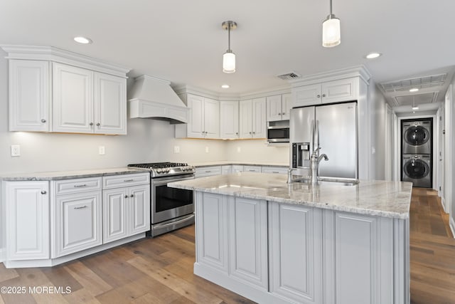 kitchen with appliances with stainless steel finishes, stacked washer / dryer, white cabinets, a sink, and premium range hood