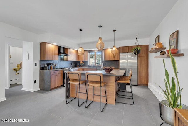 kitchen featuring a baseboard heating unit, brown cabinets, a kitchen breakfast bar, stainless steel appliances, and wall chimney exhaust hood